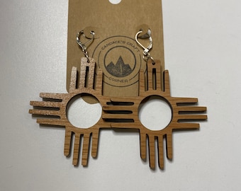 Peut inclure: Une paire de boucles d'oreilles en bois représentant le symbole du soleil Zia, un symbole traditionnel du peuple Pueblo du Nouveau-Mexique. Les boucles d'oreilles sont brunes et ont un crochet argenté.