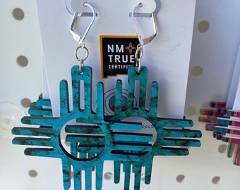 Peut inclure: Une paire de boucles d'oreilles bleu turquoise avec un motif du Sud-Ouest. Les boucles d'oreilles sont en bois et ont un crochet argenté. Les boucles d'oreilles sont étiquetées "NM True Certified".