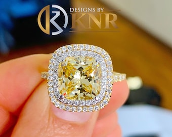 May include: A yellow cushion-cut diamond engagement ring with a double halo setting. The ring is set in yellow gold and features a row of smaller diamonds around the center stone.