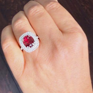 Może przedstawiać: Duży pierścionek z rubinem w owalnym kształcie z otoczką z małych, błyszczących diamentów. Rubin ma głęboki czerwony kolor, a diamenty są czyste i jasne. Pierścionek osadzony jest w srebrnej oprawie.