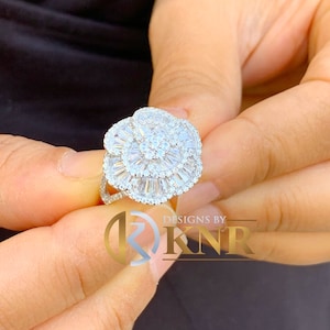 Puede incluir: Un anillo de plata con un gran diseño en forma de flor engastado con muchos pequeños diamantes transparentes. El anillo está en un dedo.