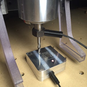 May include: A close-up of a CNC machine drilling a hole in a metal plate with a red light on the plate. The plate has a hexagonal design with a logo in the center.