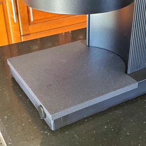 May include: A black, rectangular base with a textured surface and a round, black knob on the side. The base is part of a larger, black appliance.