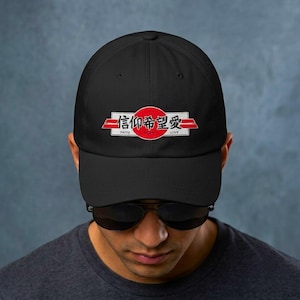 Peut inclure: Une casquette de baseball noire ornée d'un motif central composé d'un cercle rouge et de bandes horizontales blanches. Le design inclut des caractères kanji japonais noirs et les mots anglais Faith, Hope et Love inscrits en blanc.