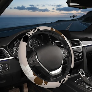 May include: Steering wheel cover with a floral pattern in black, white, and brown. The cover is beige and is installed in a car with a view of the ocean and a road.