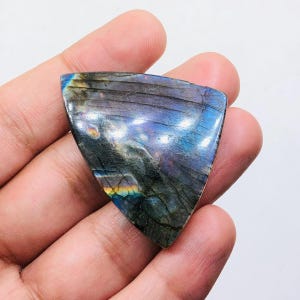 Può includere: Una pietra preziosa di labradorite lucidata, triangolare, con riflessi iridescenti blu, viola e verdi. La pietra ha un colore di base grigio scuro con striature visibili e una superficie liscia e riflettente. La pietra è tenuta in una mano.