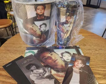 May include: A set of merchandise featuring a K-pop group. Includes a black tumbler and a silver cup with images of the group members. Also, several photo cards are visible, with the text "I AM STILL" on one.