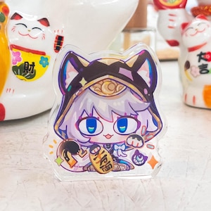 Könnte beinhalten: Ein klarer Acryl-Charm mit einer Cartoonfigur mit blauen Augen, einem goldenen Kopfschmuck und einem lila-schwarzen Outfit. Der Charm befindet sich vor mehreren weißen Keramik-Maneki-neko-Glückskatzenfiguren.