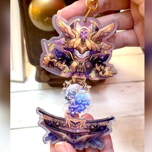 以下が含まれることがあります： 羽根の生えた金色のキャラクターが指揮棒を持っている、透明なアクリル製のキーホルダー。キャラクターは、ガラスの球体の中に小さな青いキャラクターがいるプラットフォームの上に立っています。キーホルダーは金色の金属チェーンが付いています。