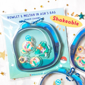 May include: Shakeable charm necklaces featuring Pokemon characters Rowlet and Meltan in a blue and white bag design. The text "Rowlet & Meltan in Ash's Bag" and "Shakeable" are printed on the card.
