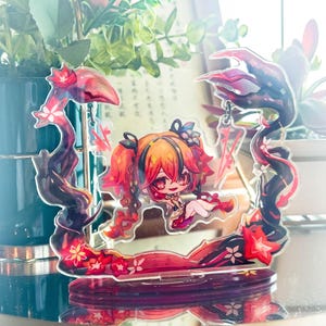 May include: A decorative acrylic figure featuring a stylized anime character with red and orange hair. The figure is set within a frame of dark, swirling lines with red floral accents. The base is a dark red color.