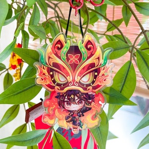 以下が含まれることがあります： ライオンダンスの衣装を着た漫画のキャラクターが描かれたカラフルなアクリルチャーム。チャームは、バネ式の留め具が付いた赤い金属製のキーホルダーに取り付けられています。