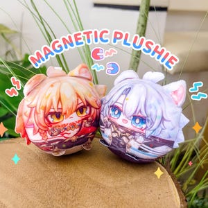 以下が含まれることがあります： アニメスタイルのキャラクターデザインの、丸いぬいぐるみ2つ。1つはオレンジと赤、もう1つは白と紫です。「MAGNETIC PLUSHIE」の文字が上にあります。ぬいぐるみは猫のような耳をしています。