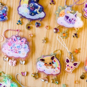 Op de afbeelding: Een verzameling kleurrijke acryl bedels met verschillende stripfiguren. Elke bedel is bevestigd aan een ketting of band en bevat decoratieve elementen zoals sterren, wolken en regenbogen. De bedels worden gepresenteerd op een lichtbruin houten oppervlak.