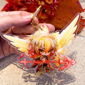 以下が含まれることがあります： 黄色い髪、翼、赤い槍を持つちびアニメキャラクターが特徴の透明なアクリルキーホルダー。キーホルダーはゴールドチェーンに付いています。赤とオレンジの装飾品が背景にあります。