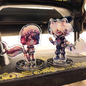 Può includere: Due figurine di personaggi in stile anime su supporti in acrilico trasparente. La figurina a sinistra è una ragazza con lunghi capelli neri, vestita con un abito bianco e nero e che tiene una spada. La figurina a destra è una creatura simile a un lupo con pelliccia bianca, vestita con un abito nero e rosso e che tiene un'arma. Entrambe le figurine hanno dettagli colorati e sono posizionate su uno sfondo nero.