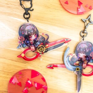 May include: Keychains featuring anime-style characters with scissors. One keychain has a cat-shaped clasp. The scissors are red and gold. The other keychain has a star-shaped clasp. The background includes red and pink circular coasters with geometric designs.