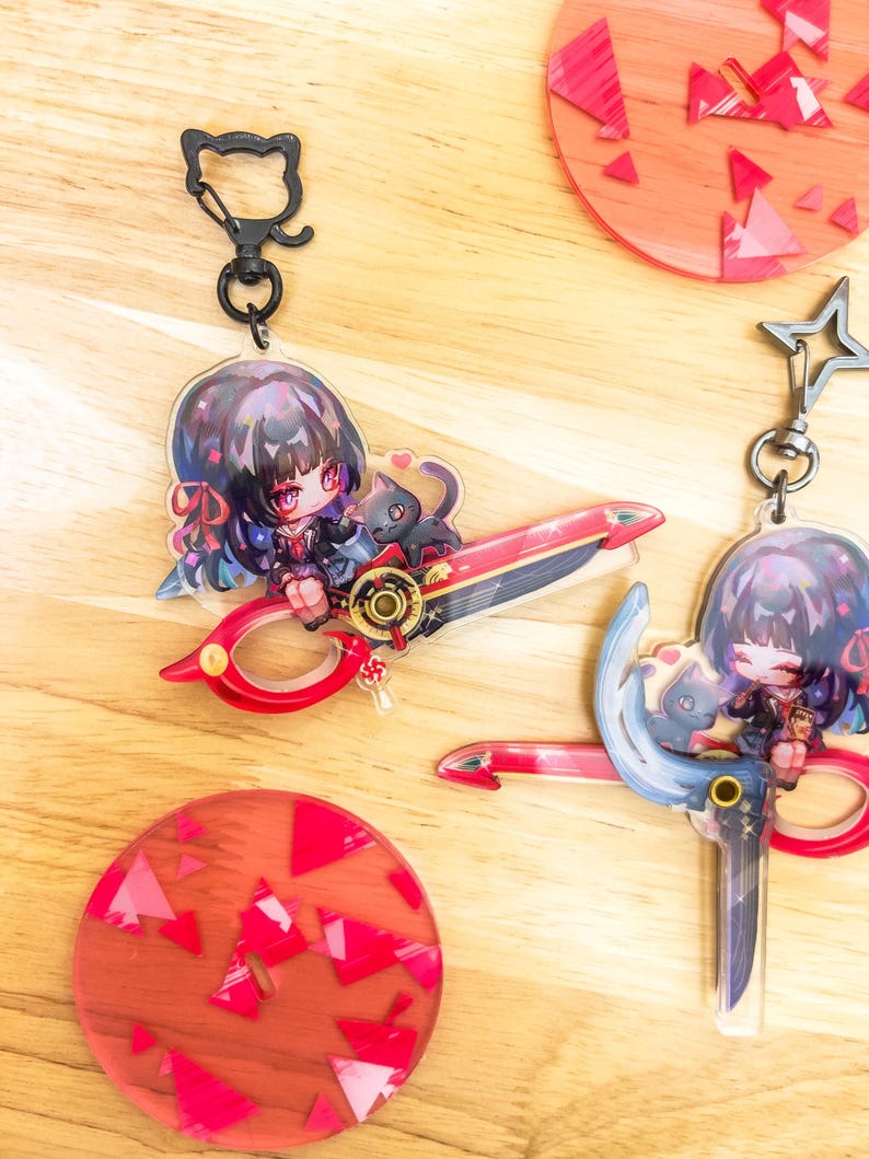 May include: Keychains featuring anime-style characters with scissors. One keychain has a cat-shaped clasp. The scissors are red and gold. The other keychain has a star-shaped clasp. The background includes red and pink circular coasters with geometric designs.