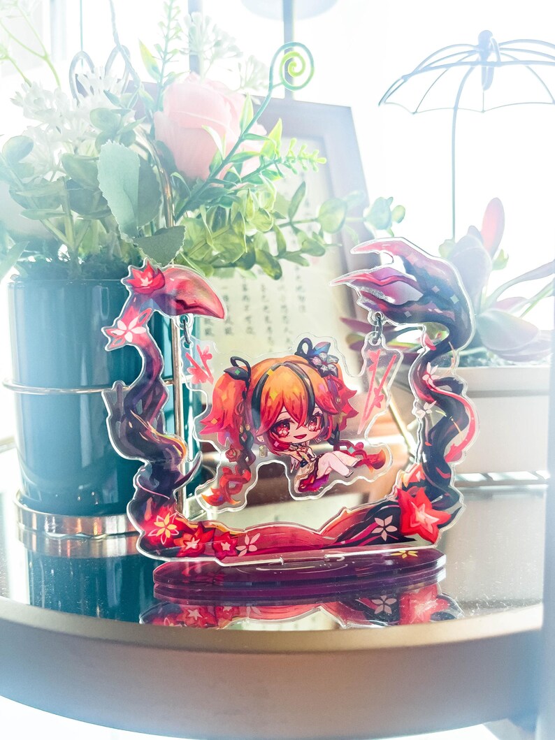 May include: A decorative acrylic figure featuring a stylized anime character with red and orange hair. The figure is set within a frame of dark, swirling lines with red floral accents. The base is a dark red color.
