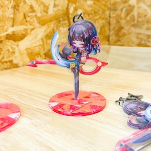 May include: A colorful anime-style keychain featuring a character with purple hair and a black cat, perched on a red and blue scissor-shaped design. The keychain is mounted on a red circular base. Additional keychains are visible.