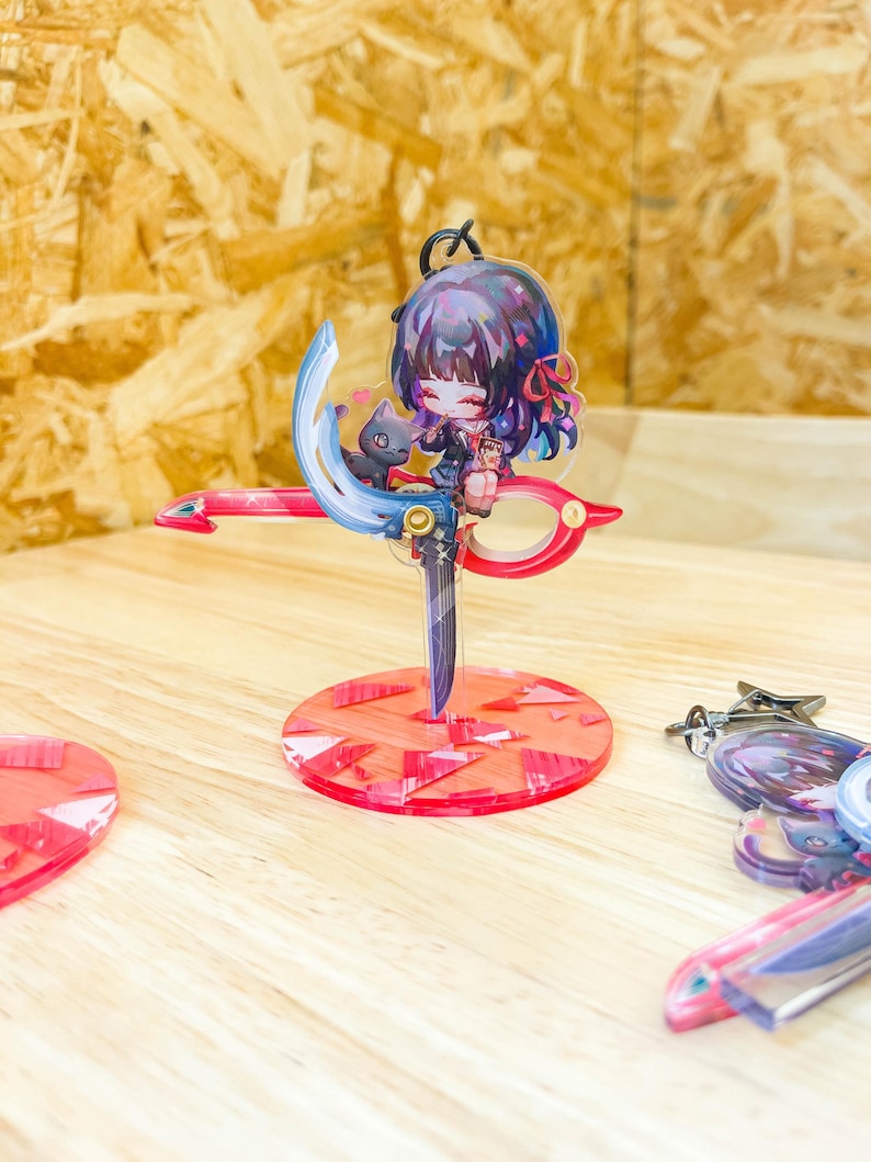 May include: A colorful anime-style keychain featuring a character with purple hair and a black cat, perched on a red and blue scissor-shaped design. The keychain is mounted on a red circular base. Additional keychains are visible.