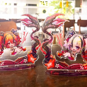 May include: Two acrylic anime-style figures featuring a chibi character with red and orange hair, and another with white and blonde hair. The figures are set on a red base with floral accents and a dark, swirling design. The figures are on a wooden table.