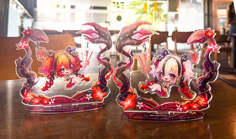 May include: Two acrylic anime-style figures featuring a chibi character with red and orange hair, and another with white and blonde hair. The figures are set on a red base with floral accents and a dark, swirling design. The figures are on a wooden table.
