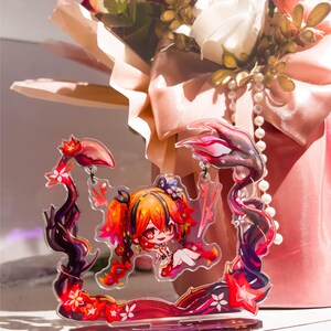 May include: A clear acrylic charm featuring a chibi anime character with orange and red hair, set within a decorative frame of dark red and black floral elements. The background includes a bouquet of red and white flowers with a pink ribbon.