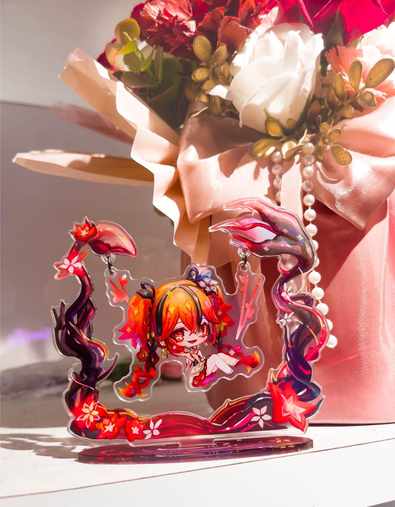 May include: A clear acrylic charm featuring a chibi anime character with orange and red hair, set within a decorative frame of dark red and black floral elements. The background includes a bouquet of red and white flowers with a pink ribbon.