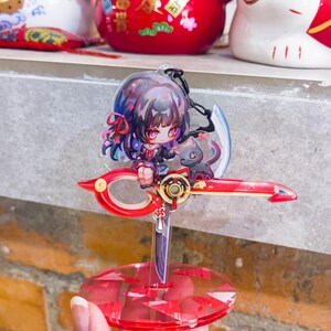 May include: A red and clear acrylic anime-style figure featuring a girl with dark hair and a black cat. The figure is mounted on a red scissor-shaped base with a clear, round stand.