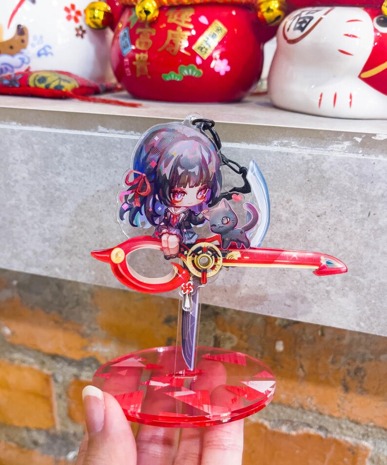 May include: A red and clear acrylic anime-style figure featuring a girl with dark hair and a black cat. The figure is mounted on a red scissor-shaped base with a clear, round stand.