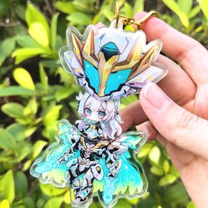 Op de afbeelding: Een transparante acryl sleutelhanger met een chibi anime-personage in een wit en gouden harnas met blauwe accenten. Het personage houdt een zwaard vast en heeft een blauw-groene cape.