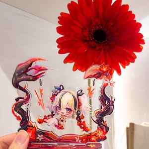 May include: A clear acrylic stand featuring a cartoon character with blonde hair and pigtails. The stand has a red and black floral design. A large red gerbera daisy is in the background.