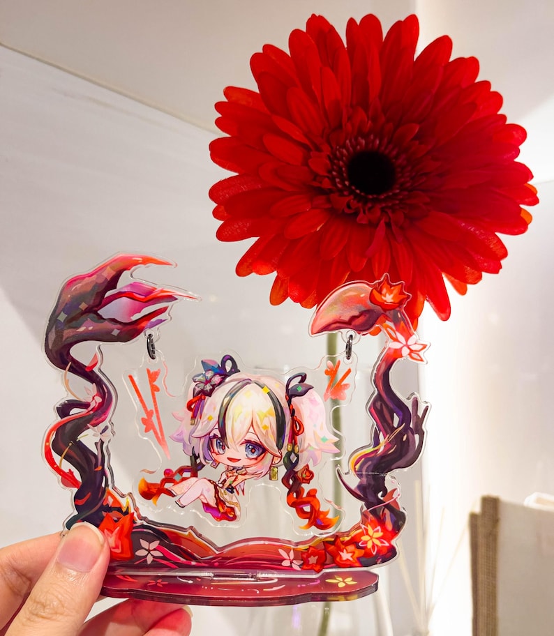 May include: A clear acrylic stand featuring a cartoon character with blonde hair and pigtails. The stand has a red and black floral design. A large red gerbera daisy is in the background.