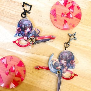 May include: Anime-style keychains featuring a character with dark hair and a black cat, holding a red and blue weapon. The keychains have a cat-shaped clasp and a star-shaped clasp. Two round coasters with red and pink geometric designs are also visible.