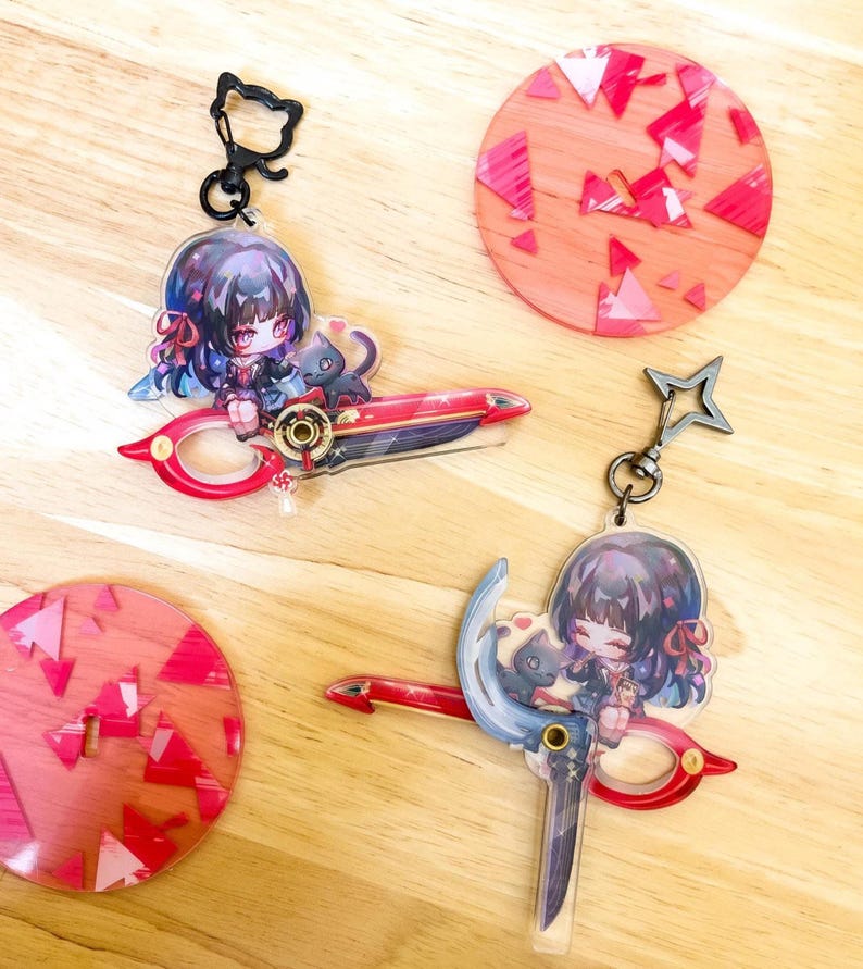 May include: Anime-style keychains featuring a character with dark hair and a black cat, holding a red and blue weapon. The keychains have a cat-shaped clasp and a star-shaped clasp. Two round coasters with red and pink geometric designs are also visible.