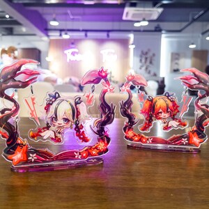 May include: Two acrylic anime character figures. Each figure features a chibi-style girl with light-colored hair and red accents. The figures are framed by a decorative border with floral and abstract elements in red and black. They are displayed on a wooden table.