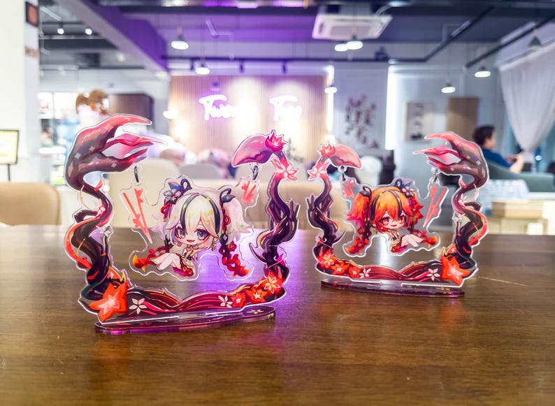 May include: Two acrylic anime character figures. Each figure features a chibi-style girl with light-colored hair and red accents. The figures are framed by a decorative border with floral and abstract elements in red and black. They are displayed on a wooden table.