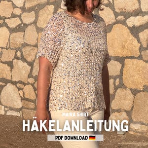 Op de afbeelding: Een beige gehaakt shirt met korte mouwen, voorzien van een gestructureerd breipatroon ingelegd met talloze reflecterende zilveren lovertjes. Het kledingstuk heeft een ronde halslijn en een rechte zoom, met de tekst MAIRA SHIRT HÄKELANLEITUNG PDF DOWNLOAD.