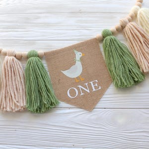 May include: A burlap banner with a cartoon duck and the word "ONE" in white letters. The banner is strung with wooden beads and green, white, and beige yarn tassels.