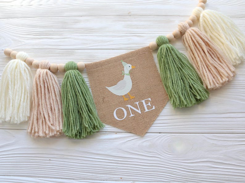 May include: A burlap banner with a cartoon duck and the word "ONE" in white letters. The banner is strung with wooden beads and green, white, and beige yarn tassels.