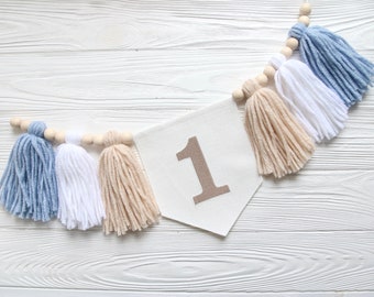 May include: A white banner with the number "1" in brown fabric, decorated with a string of wooden beads and blue, white, and beige yarn tassels.