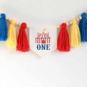 May include: A white banner with a red and white striped circus tent and the word "ONE" in blue. The banner is strung with wooden beads and yellow, red, and blue yarn tassels.
