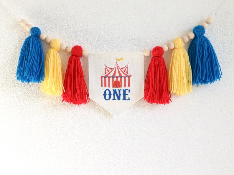 May include: A white banner with a red and white striped circus tent and the word "ONE" in blue. The banner is strung with wooden beads and yellow, red, and blue yarn tassels.