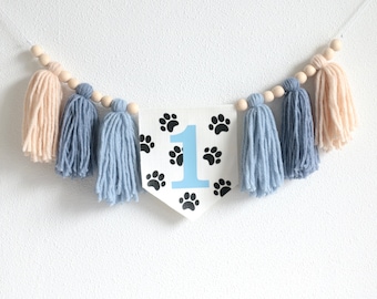 May include: A white banner with black paw prints and a blue number one. The banner is strung with wooden beads and blue and beige yarn tassels.