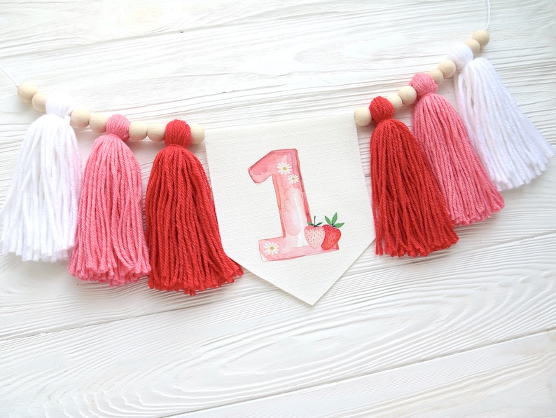 Puede incluir: Una guirnalda de borlas blancas, rosas y rojas con una pancarta blanca que dice "1" en rosa con fresas y flores.