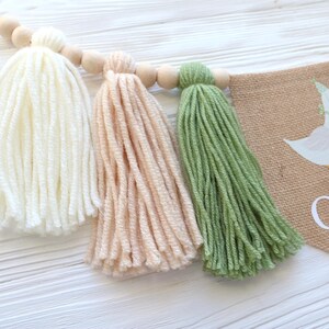 May include: A burlap banner with a white goose illustration and the text "One" in white. The banner is decorated with three yarn tassels in white, peach, and green.