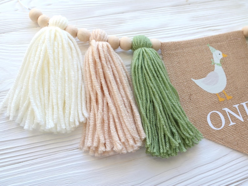 May include: A burlap banner with a white goose illustration and the text "One" in white. The banner is decorated with three yarn tassels in white, peach, and green.