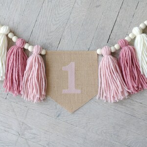May include: A burlap banner with the number "1" in pink, decorated with pink and white yarn tassels and wooden beads.