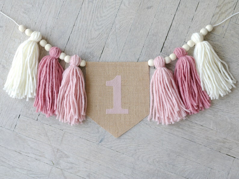 May include: A burlap banner with the number "1" in pink, decorated with pink and white yarn tassels and wooden beads.